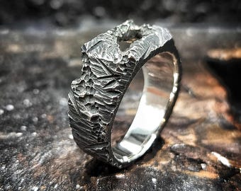 May include: A textured silver ring with a rugged, organic design. The ring features a rough, uneven surface, creating a unique, handcrafted appearance. The band is approximately 0.5 inches wide, with a polished interior.