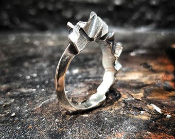 Può includere: Un anello d'argento con un design geometrico e angolare. L'anello presenta una serie di forme appuntite e sfaccettate che creano un aspetto a forma di corona. L'anello è posto su uno sfondo scuro e strutturato.