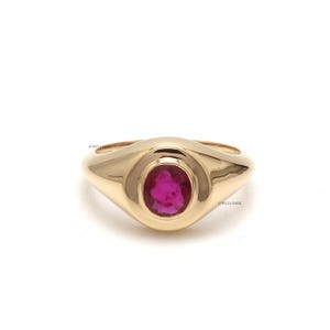 Puede incluir: Un anillo de oro con una gran gema ovalada de color rubí en el centro. El anillo tiene un acabado liso y pulido y un diseño clásico y minimalista. El anillo es una joya.