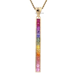 Op de afbeelding: Een gouden ketting met een verticale hanger die is bezet met een regenboog van vierkante edelstenen. De edelstenen zijn in een verticale lijn gerangschikt, beginnend met rood aan de bovenkant en eindigend met geel aan de onderkant. De hanger is bevestigd aan een gouden ketting.