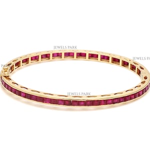 Pode incluir: Um bracelete de ouro com uma fila de pedras preciosas vermelhas quadradas. O bracelete é aberto e tem um fecho com dobradiça.