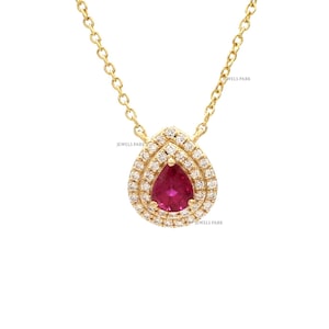 Puede incluir: Un collar de oro con un colgante en forma de pera. El colgante presenta una piedra preciosa roja vibrante en el centro, rodeada por un halo de piedras más pequeñas y brillantes. La delicada cadena y el colgante son de un tono dorado cálido.