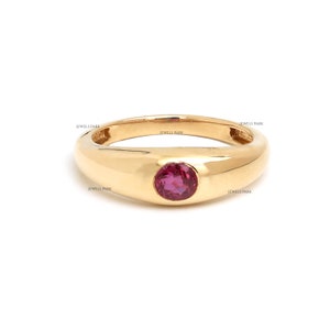 Puede incluir: Un anillo de oro con una sola piedra preciosa roja redonda engastada en el centro. El anillo tiene un diseño simple y clásico.