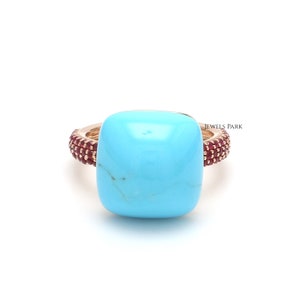 Op de afbeelding: Een ring met een vierkante turquoise steen, gezet in een gouden band. De band is versierd met kleine rode edelstenen.