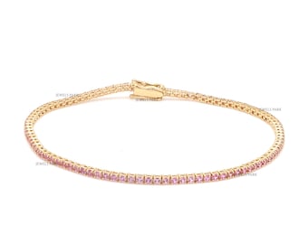May include: A gold tennis bracelet with pink gemstones. The bracelet is made of a delicate chain with small, square-shaped gemstones set along the edge.