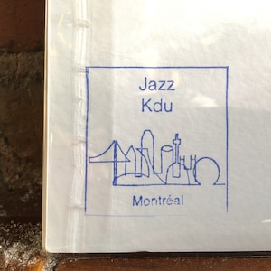 K&ouml;nnte beinhalten: Ein wei&szlig;es Papier mit einer blau getuschten Illustration der Skyline von Montreal, einschlie&szlig;lich einer Br&uuml;cke und verschiedener Geb&auml;ude. Der Text "Jazz Kdu Montr&eacute;al" ist in Blau in einem quadratischen Rahmen gedruckt.