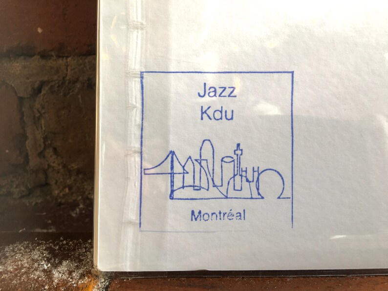 K&ouml;nnte beinhalten: Ein wei&szlig;es Papier mit einer blau getuschten Illustration der Skyline von Montreal, einschlie&szlig;lich einer Br&uuml;cke und verschiedener Geb&auml;ude. Der Text "Jazz Kdu Montr&eacute;al" ist in Blau in einem quadratischen Rahmen gedruckt.