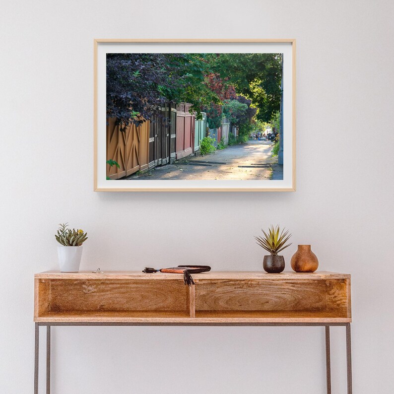 K&ouml;nnte beinhalten: Gerahmtes Foto einer sonnendurchfluteten Gasse mit bunten Holzz&auml;unen und &uuml;ppigem Gr&uuml;n. Das Bild f&auml;ngt ein Gef&uuml;hl von Tiefe und Ruhe ein, wobei das Sonnenlicht Schatten auf den B&uuml;rgersteig wirft. Das Foto wird auf einer Holzkonsolentisch pr&auml;sentiert.