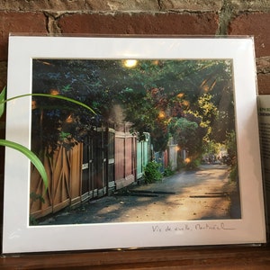 K&ouml;nnte beinhalten: Ein gerahmter Druck einer Stra&szlig;enszene in Montreal mit einer Reihe farbenfroher Holzz&auml;une und &uuml;ppigem Gr&uuml;n. Der Druck ist signiert mit "Vic de aveille, Montr&eacute;al". Das Bild befindet sich in einem wei&szlig;en Passepartout.