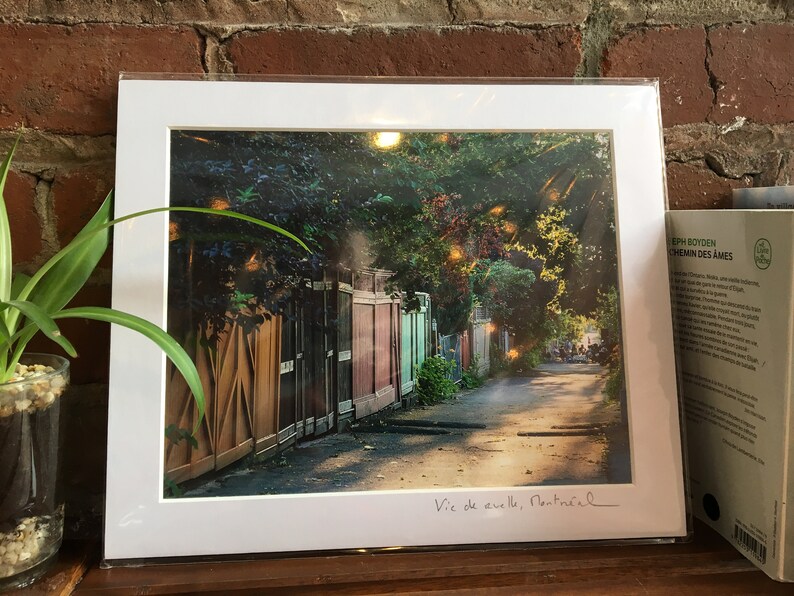 K&ouml;nnte beinhalten: Ein gerahmter Druck einer Stra&szlig;enszene in Montreal mit einer Reihe farbenfroher Holzz&auml;une und &uuml;ppigem Gr&uuml;n. Der Druck ist signiert mit "Vic de aveille, Montr&eacute;al". Das Bild befindet sich in einem wei&szlig;en Passepartout.