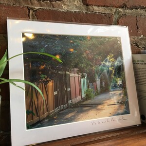 K&ouml;nnte beinhalten: Ein gerahmter Druck einer Stra&szlig;enszene in Montreal mit einer Reihe farbenfroher Holzz&auml;une entlang eines Weges. Der Druck ist wei&szlig; gerahmt und tr&auml;gt den handgeschriebenen Text "Vie de aville, Montreal". Im Hintergrund ist ein Buch zu sehen.