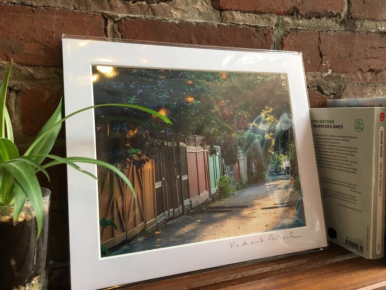 K&ouml;nnte beinhalten: Ein gerahmter Druck einer Stra&szlig;enszene in Montreal mit einer Reihe farbenfroher Holzz&auml;une entlang eines Weges. Der Druck ist wei&szlig; gerahmt und tr&auml;gt den handgeschriebenen Text "Vie de aville, Montreal". Im Hintergrund ist ein Buch zu sehen.