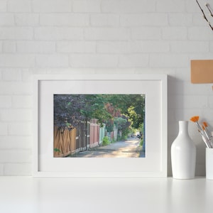 K&ouml;nnte beinhalten: Ein gerahmtes Foto eines sonnigen Weges, ges&auml;umt von bunten Holzz&auml;unen und &uuml;ppigem Gr&uuml;n. Das Bild wird auf einer wei&szlig;en Oberfl&auml;che pr&auml;sentiert, mit einer wei&szlig;en Backsteinmauer im Hintergrund. Dekorative Elemente wie Vasen und Pinsel sind ebenfalls vorhanden.