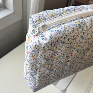 May include: A quilted floral print cosmetic bag with a white zipper and a heart-shaped zipper pull. The bag features a pattern of small yellow, blue, and pink flowers on a white background. The bag is rectangular and appears to be made of soft fabric.