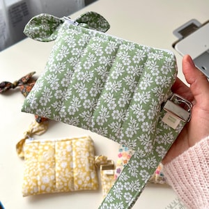 Puede incluir: Un estuche acolchado de color verde salvia con un estampado floral blanco, cierre de cremallera superior y una correa de tela a juego con una hebilla de metal plateado. Una peque&ntilde;a etiqueta blanca muestra el texto HAND MADE.