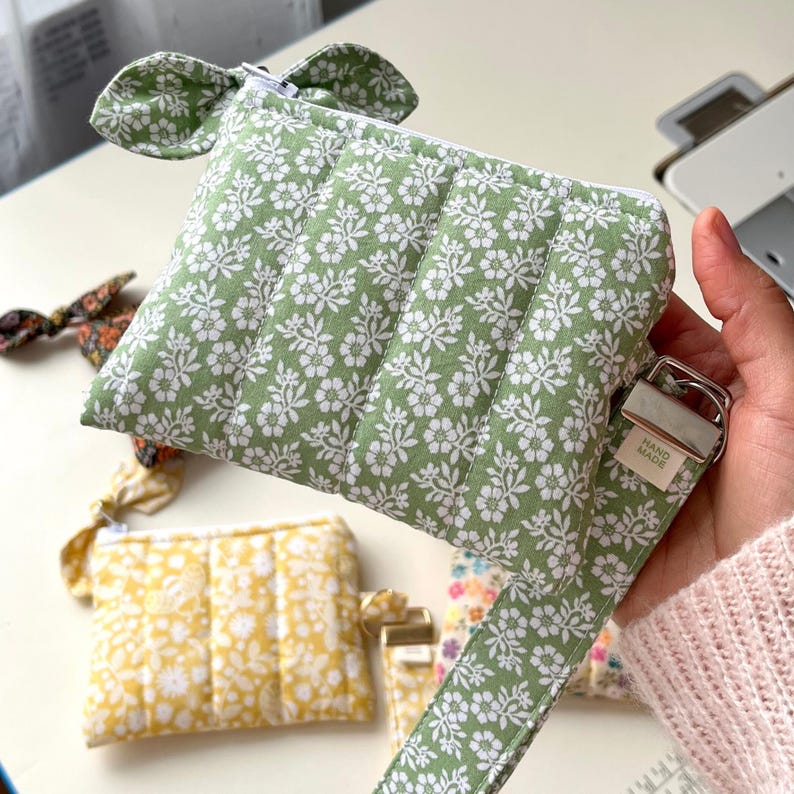 Puede incluir: Un estuche acolchado de color verde salvia con un estampado floral blanco, cierre de cremallera superior y una correa de tela a juego con una hebilla de metal plateado. Una peque&ntilde;a etiqueta blanca muestra el texto HAND MADE.