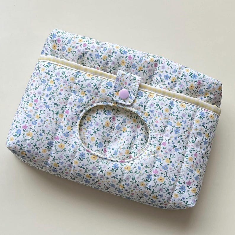 May include: A quilted floral print tissue holder with a light yellow trim and a button closure. The fabric features a pattern of small blue, yellow, and purple flowers on a white background. The holder has an oval opening for easy access to tissues.