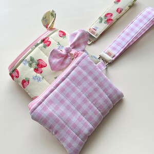 Puede incluir: Dos bolsos de tela acolchada con cierre de cremallera. Uno tiene un fondo crema con fresas rojas y flores azules, mientras que el otro presenta un patr&oacute;n de cuadros vichy rosa y blanco. Ambos incluyen correas de mu&ntilde;eca con etiquetas.