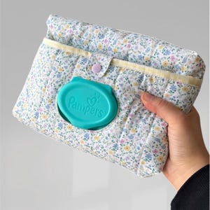 May include: A floral print baby wipes case with a teal lid that reads "Pampers". The case is quilted with a pastel floral pattern and a yellow trim. It is held in a person's hand.
