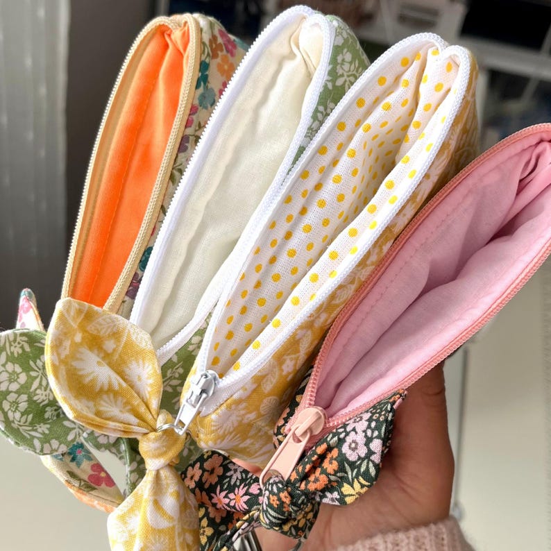 Puede incluir: Un conjunto de cuatro estuches de tela con cremallera que presentan varios estampados florales y de lunares en tonos naranja, amarillo, verde y rosa. Cada estuche tiene un lazo de tela a juego en el tirador de la cremallera y un forro liso.