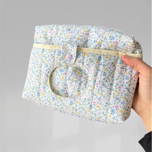 May include: A floral-patterned quilted pouch with a light blue, yellow, and pink flower design. The pouch has a circular opening and a button closure. The background is a solid light gray.