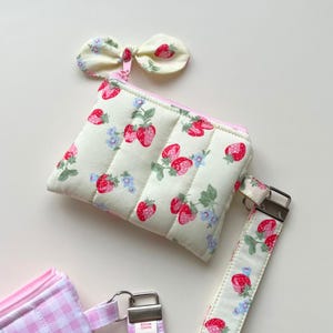 Puede incluir: Un estuche de tela acolchada con fondo crema y estampado de fresas rojas y flores azules. El estuche tiene una cremallera rosa, un lazo decorativo de tela en el tirador y una correa de mu&ntilde;eca a juego con herrajes de color plateado.