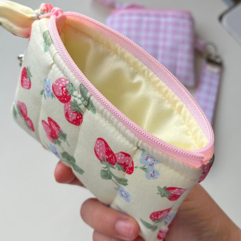 Puede incluir: Un peque&ntilde;o estuche acolchado de tela color crema con un patr&oacute;n repetitivo de fresas rojas y peque&ntilde;as flores azules. El estuche tiene una cremallera de color rosa claro, un forro interior amarillo p&aacute;lido y una correa a juego.
