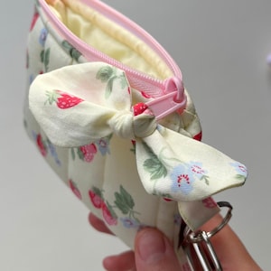 Puede incluir: Un peque&ntilde;o estuche de tela color crema con un patr&oacute;n repetitivo de fresas rojas y peque&ntilde;as flores azules. El estuche tiene una cremallera rosa claro, un lazo de tela decorativo en el lateral y una anilla de metal plateado.