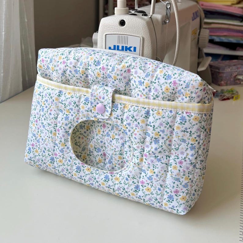 May include: A floral print tissue box cover with a button closure. The cover is white with a pattern of small blue, yellow, and purple flowers and green leaves. The edges are trimmed with yellow fabric. The cover has a large oval opening.