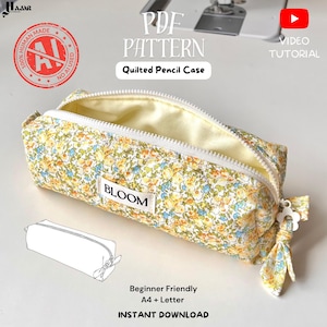 May include: A quilted pencil case featuring a yellow, blue, and green floral pattern with a white zipper and a white fabric label reading BLOOM. The case includes a white flower-shaped zipper pull and a matching floral fabric tie.
