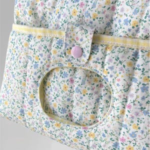 May include: Close-up of a quilted fabric bag with a floral pattern in pastel colors. The bag features a large oval opening, a yellow trim, and a pink button closure. The fabric is white with blue, yellow, and pink flowers.