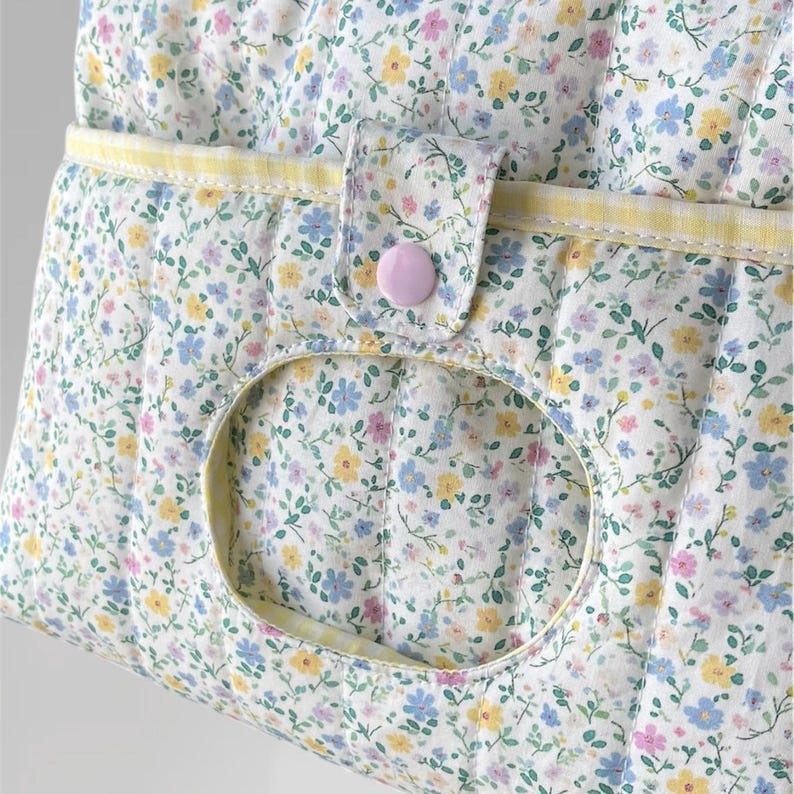 May include: Close-up of a quilted fabric bag with a floral pattern in pastel colors. The bag features a large oval opening, a yellow trim, and a pink button closure. The fabric is white with blue, yellow, and pink flowers.