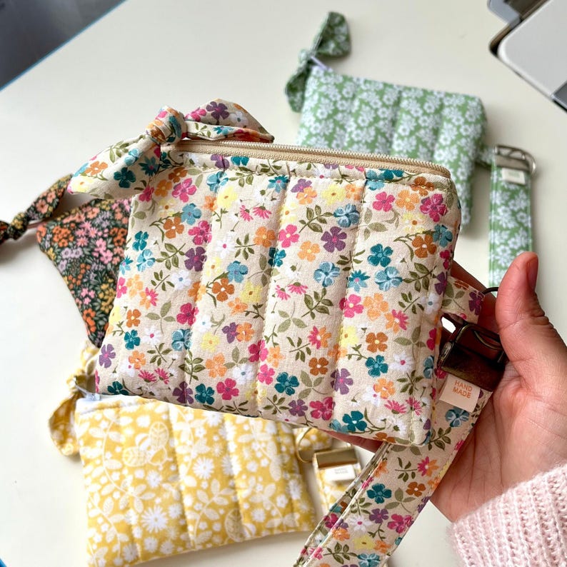 Puede incluir: Un estuche de tela acolchada con un estampado floral colorido sobre fondo crema y una correa de mu&ntilde;eca a juego. El estuche tiene cierre de cremallera, un lazo de tela decorativo en el lateral y una etiqueta que dice Hand Made.