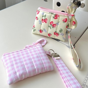 Puede incluir: Dos estuches acolchados con cremallera. Uno tiene un fondo crema con estampado de fresas rojas y flores azules, cremallera rosa y correa de mu&ntilde;eca a juego. El segundo estuche tiene un patr&oacute;n de cuadros vichy rosa y blanco con una correa.