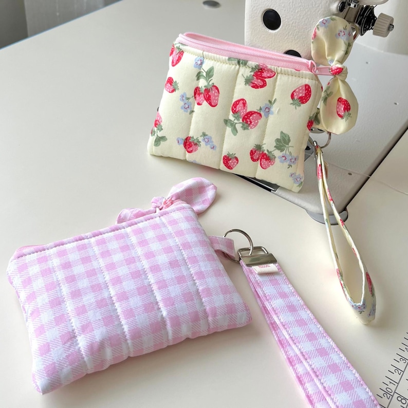 Puede incluir: Dos estuches acolchados con cremallera. Uno tiene un fondo crema con estampado de fresas rojas y flores azules, cremallera rosa y correa de mu&ntilde;eca a juego. El segundo estuche tiene un patr&oacute;n de cuadros vichy rosa y blanco con una correa.