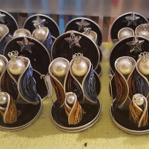 Puede incluir: Una colecci&oacute;n de doce peque&ntilde;os adornos de bel&eacute;n hechos a mano. Cada adorno presenta una base negra con un borde plateado, una figura dorada, azul y marr&oacute;n de Mar&iacute;a, Jos&eacute; y el ni&ntilde;o Jes&uacute;s, y una perla blanca para la cabeza de cada figura.