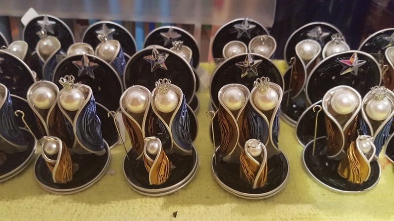 Puede incluir: Una colecci&oacute;n de doce peque&ntilde;os adornos de bel&eacute;n hechos a mano. Cada adorno presenta una base negra con un borde plateado, una figura dorada, azul y marr&oacute;n de Mar&iacute;a, Jos&eacute; y el ni&ntilde;o Jes&uacute;s, y una perla blanca para la cabeza de cada figura.
