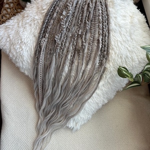 May include: A set of gray and silver dreadlocks with a wavy texture. The dreadlocks are arranged in a bundle and are adorned with small silver beads. The hair extensions are displayed on a white fluffy pillow.