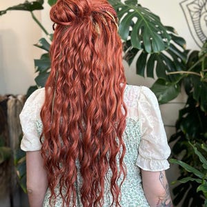 May include: Rear view of long, wavy, auburn hair styled in a bun with decorative braids. The hair cascades down over a green floral dress with white lace sleeves. The background includes lush green plants.