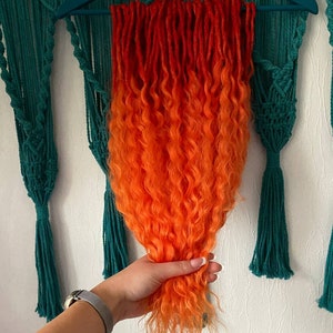 May include: Orange and red ombre curly synthetic dreadlocks hanging on a hanger. The dreadlocks are about 20 inches long.