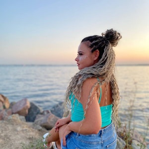May include: A woman with long, blonde, braided hair sits on a rock by the water. She is wearing a turquoise tank top and denim shorts. The sky is a light blue and the water is a calm, light blue.