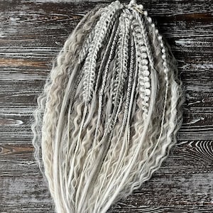 May include: A collection of gray and white synthetic dreadlocks and braids. The hair extensions feature a mix of textures, including wavy and braided sections, with a blend of light and dark gray and white tones. The hair is arranged on a dark wood surface.