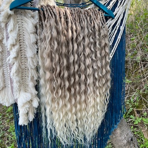 May include: A set of long, wavy, blonde and light brown synthetic dreadlocks hanging on a teal hanger. The dreadlocks are arranged in a thick, cascading waterfall style.