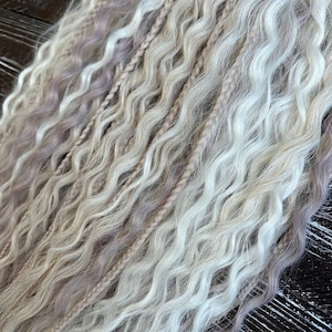 60 DE の長いウェーブヘアエクステンション。ブロンドとライトトープの色合いで、ゆるやかなテクスチャのウェーブに、細い編み込みがいくつも織り交ぜられています。