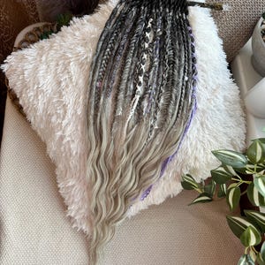May include: A set of gray, black, and purple dreadlocks with wavy ends. The dreadlocks are arranged on a fluffy white pillow. Some dreadlocks have decorative beads and white accents. The dreadlocks are held together by a dark wooden bar.
