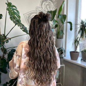 May include: A person with long, wavy brown hair styled in a half-up bun. The hair cascades down the back, with lighter highlights. The person is wearing a tie-dye style sweatshirt in shades of beige and brown. The background includes plants and a decorative wall design.