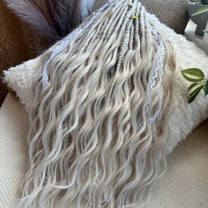 May include: A set of wavy, light blonde dreadlocks with white lace accents and gold-colored beads. The dreadlocks are arranged on a fluffy, white pillow, creating a soft and textured appearance. The hair extensions are a light, neutral color.
