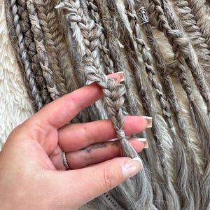 May include: A close-up of a hand holding a set of gray and white braided hair extensions. The extensions are tied together at the end and have a silver bead charm.