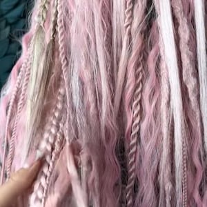 May include: Close-up of pink and white dreadlocks with various textures, including braids, twists, and wavy strands. The hair extensions are a mix of light and darker pink shades, with some white strands interspersed throughout. The overall style is bohemian and artistic.