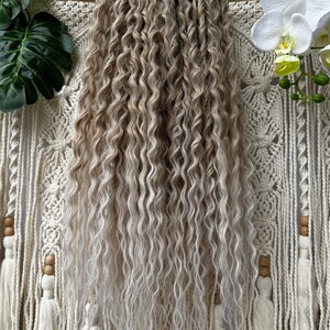 Op de afbeelding: Een bundel lange, golvende hairextensions met een lichtbruin tot blond ombre-effect. Het haar is gestyled in losse, volumineuze krullen. De extensions worden gepresenteerd tegen een crèmekleurige macramé wandhanger met houten accenten en decoratieve kwasten.